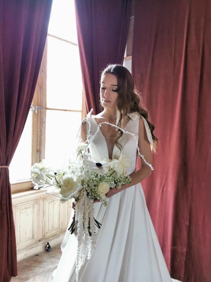 Mariée au bouquet de fleurs blanches près d’une fenêtre du château de Motteux, rideaux pourpres