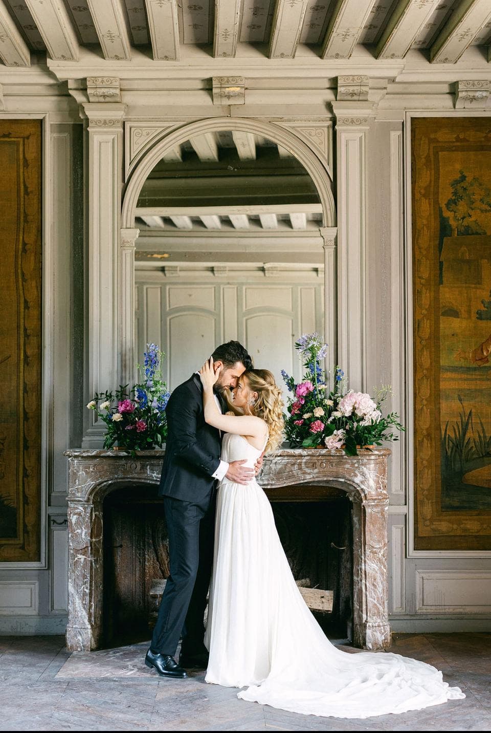 Couple de mariés enlacés devant la cheminée en marbre d’un salon historique du château de Motteux