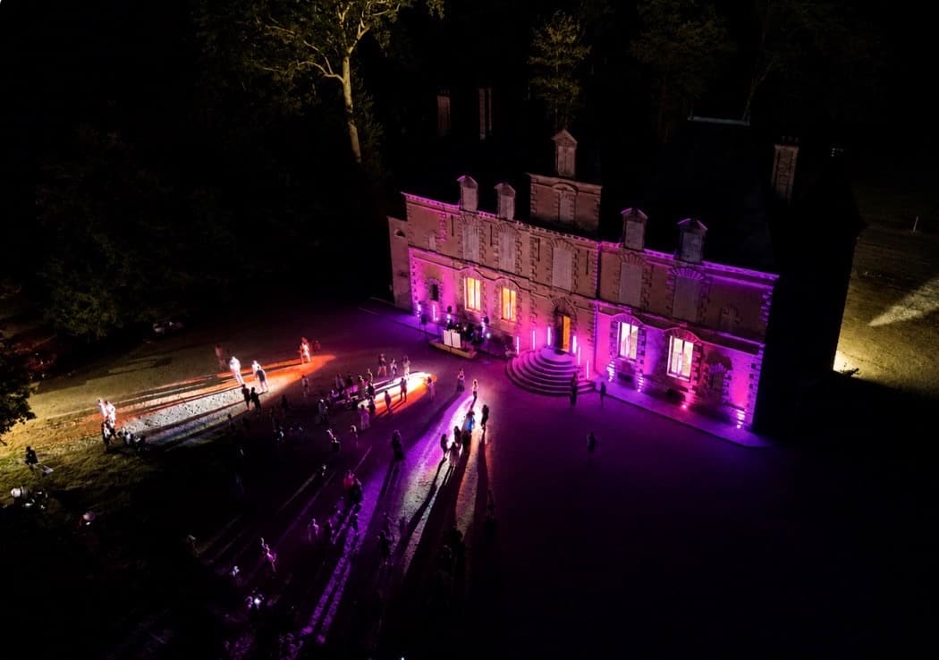 Vue aérienne nocturne du château de Motteux illuminé de rose lors d’une soirée guinguette, public sur le parvis