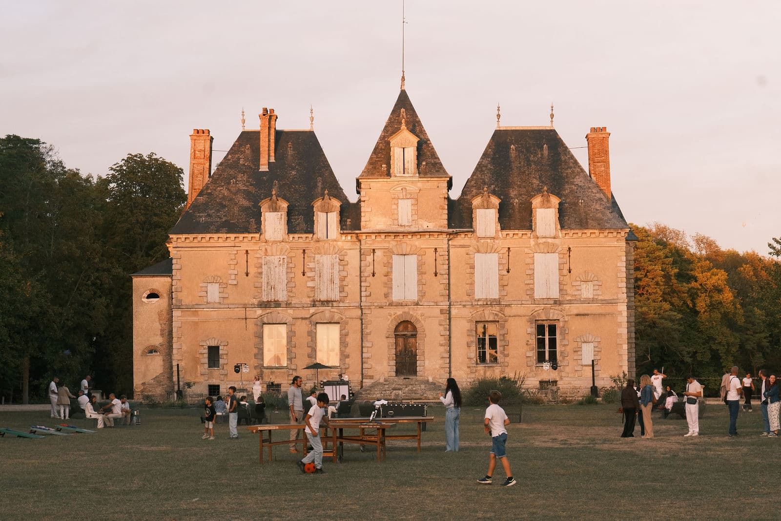Façade du château de Motteux au soleil couchant, invités vêtus de blanc et jeux sur la grande pelouse