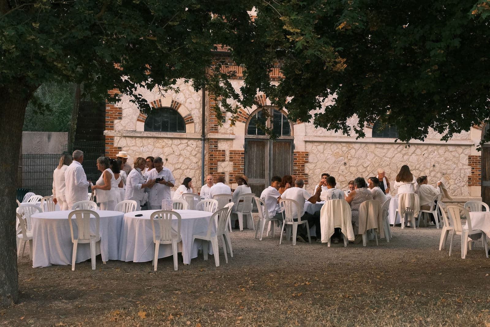 Tables rondes nappées de blanc dressées sous les arbres, le long des communs du château de Motteux