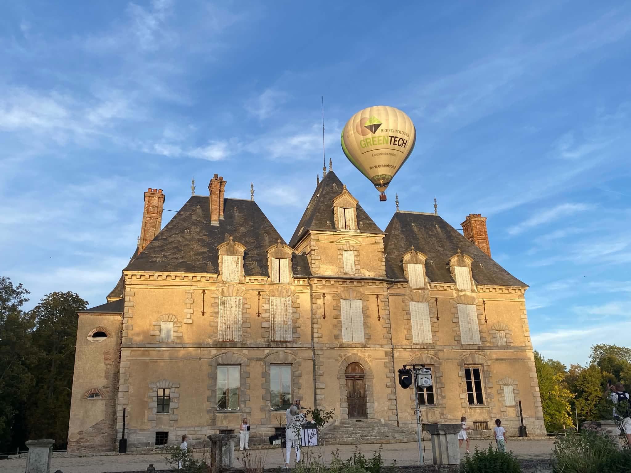 Montgolfière au-dessus de la façade du château de Motteux au soleil couchant, invités en blanc sur le parvis